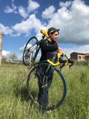 Adéla Šípová v krajině se svým kolem přes rameno. Na hlavě má cyklistickou čepici a je sportovně oblečná.